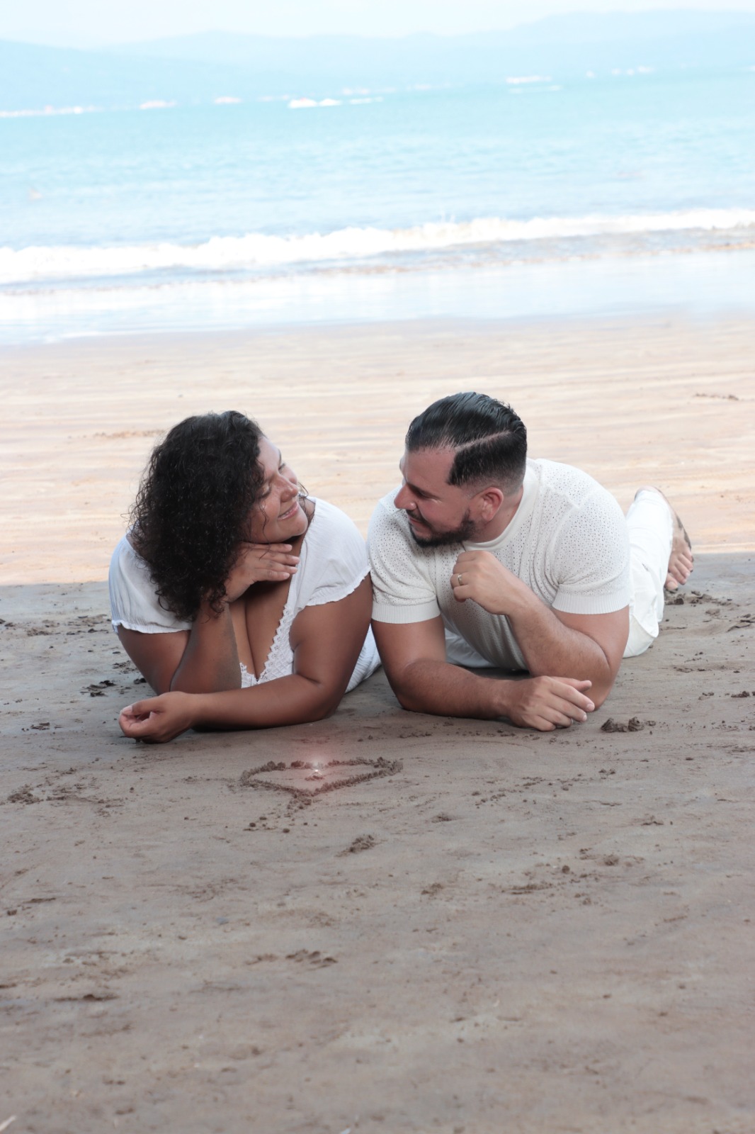 Cinthya y Adriel recostados en la arena mirándose junto a un corazón
